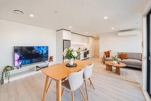 A modern living room with a flat-screen TV, a dining area with a wooden table and chairs, a sofa, and a kitchen area.