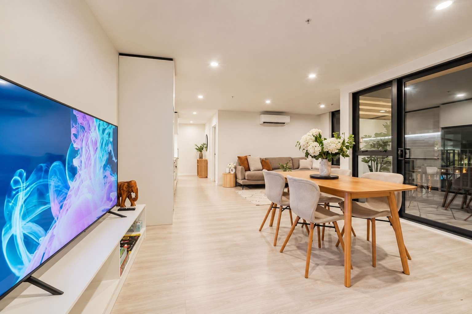 Una sala de estar moderna con un televisor de pantalla plana, un comedor con mesa y sillas de madera, y un sofá con cojines.