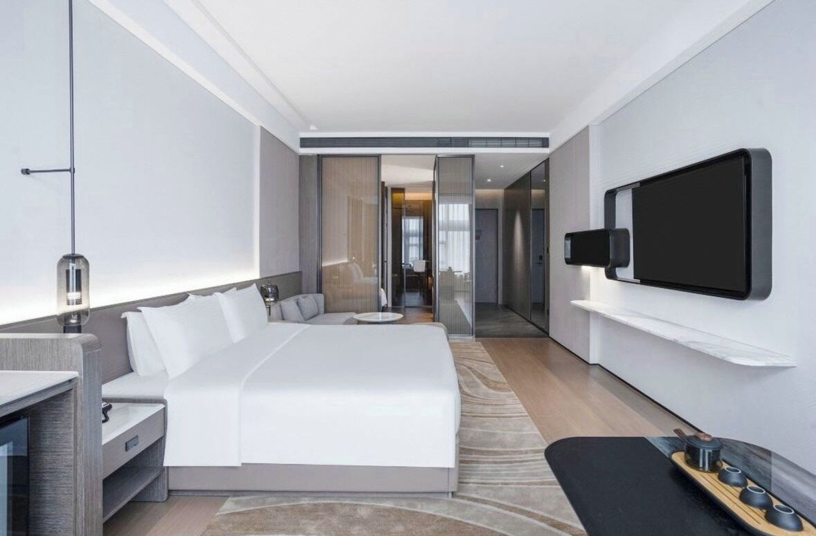 A modern hotel room with a large bed, a TV mounted on the wall, and a bedside table with a lamp.