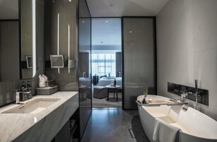 Un baño moderno con una bañera grande, un lavamanos doble y un espejo amplio. Hay un separador de vidrio que divide el baño de una sala de estar con un sofá y una mesa de centro.