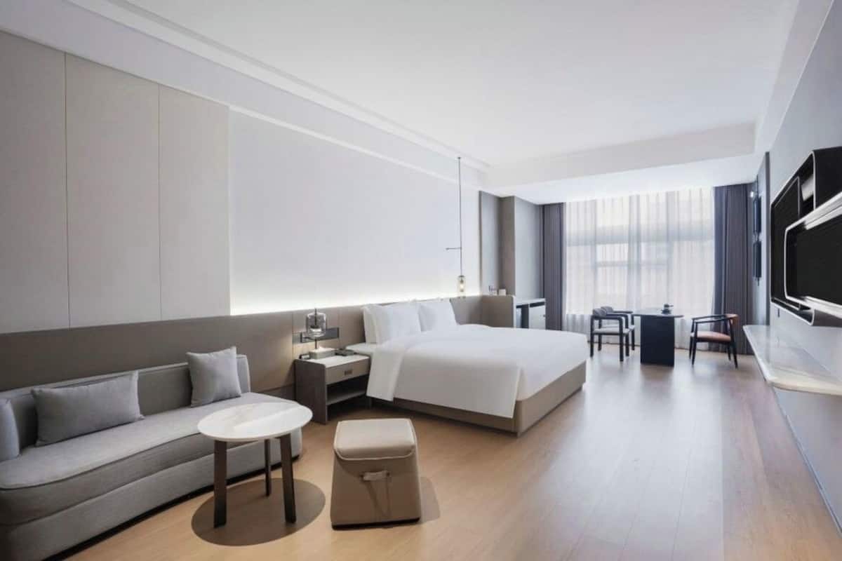Una habitación de hotel moderna con una cama grande, un sofá, una mesita, una mesita de noche, un televisor y una ventana con cortinas.