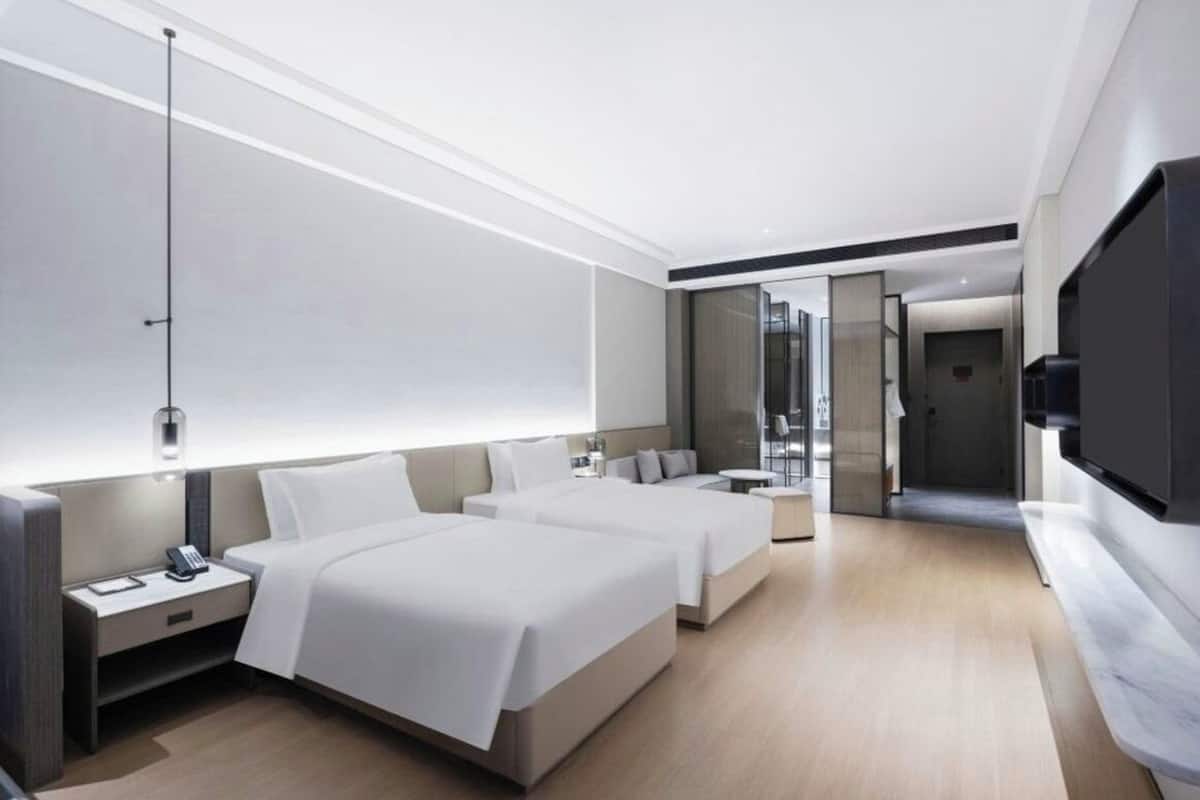 Una habitación de hotel moderna con dos camas, una mesita de noche con teléfono, un espejo grande y un televisor montado en la pared.
