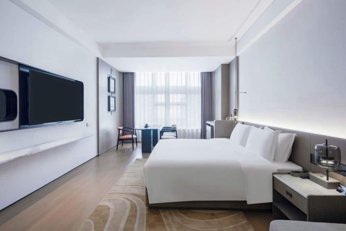 Habitación de hotel moderna con una cama grande, un escritorio, una silla, un televisor y un ventanal con cortinas.