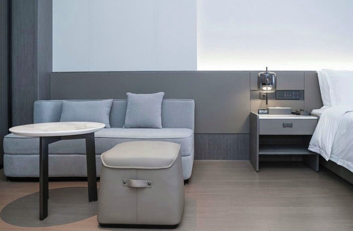Habitación de hotel moderna con un sofá gris, una mesita redonda pequeña, una mesita de noche con lámpara y una cama con ropa de cama blanca.