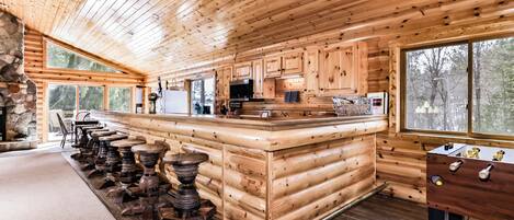 A log cabin interior with a central island bar, stone fireplace, and large windows offering views of the forest.