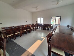 A large, rectangular conference room with a high ceiling, multiple wooden tables and chairs, and large windows allowing natural light. - Orchard Hills Resorts (Pushkar)
