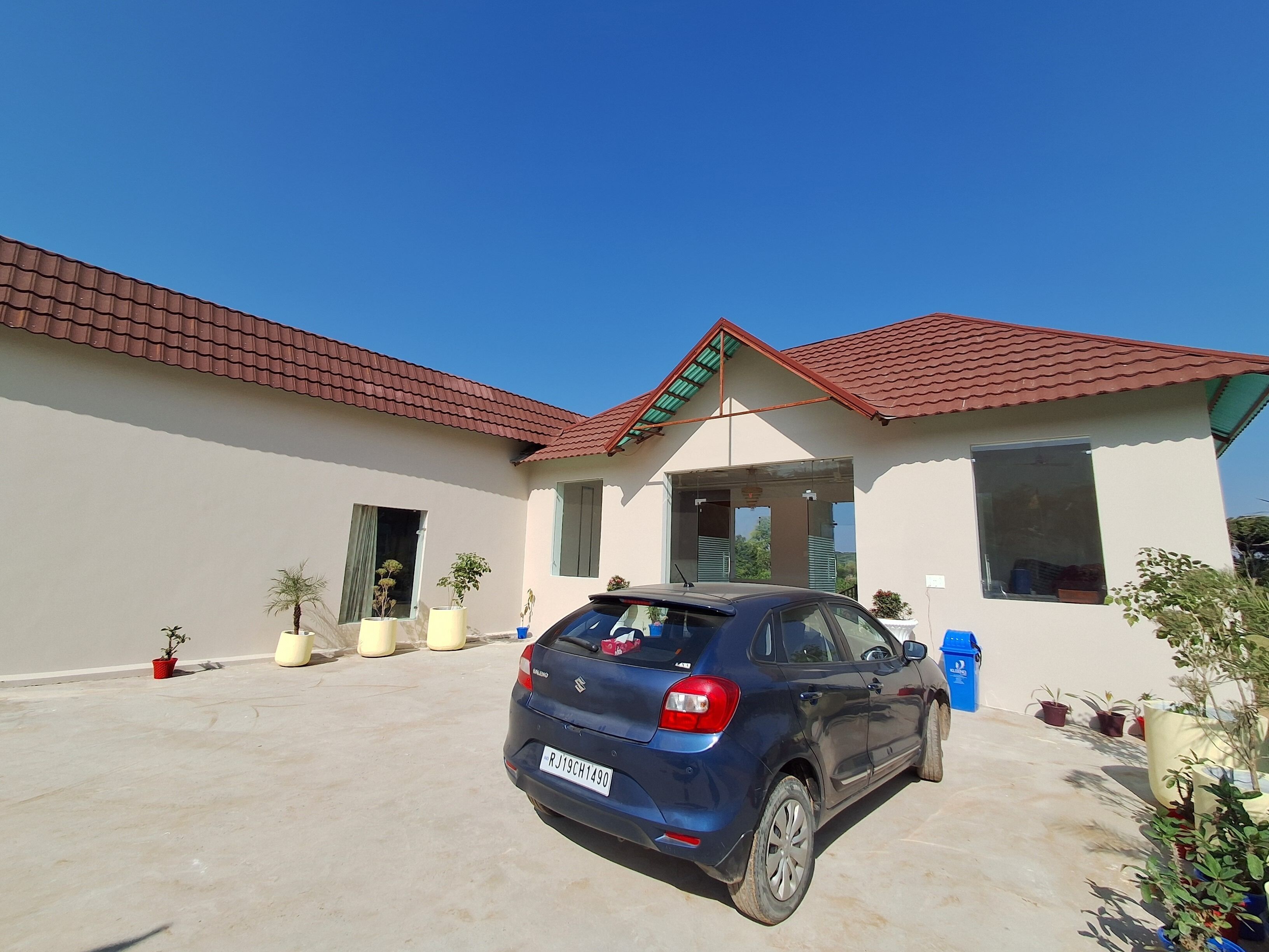 Une maison moderne avec un toit recouvert de tuiles rouges, une voiture bleue garée dans l’allée, et plusieurs plantes en pot disposées autour de la propriété.