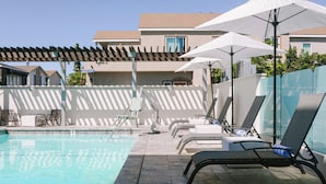 A pool area with lounge chairs and umbrellas, surrounded by a building with a tiled roof and a pergola. - Solarena at Newport Beach (Newport Beach)