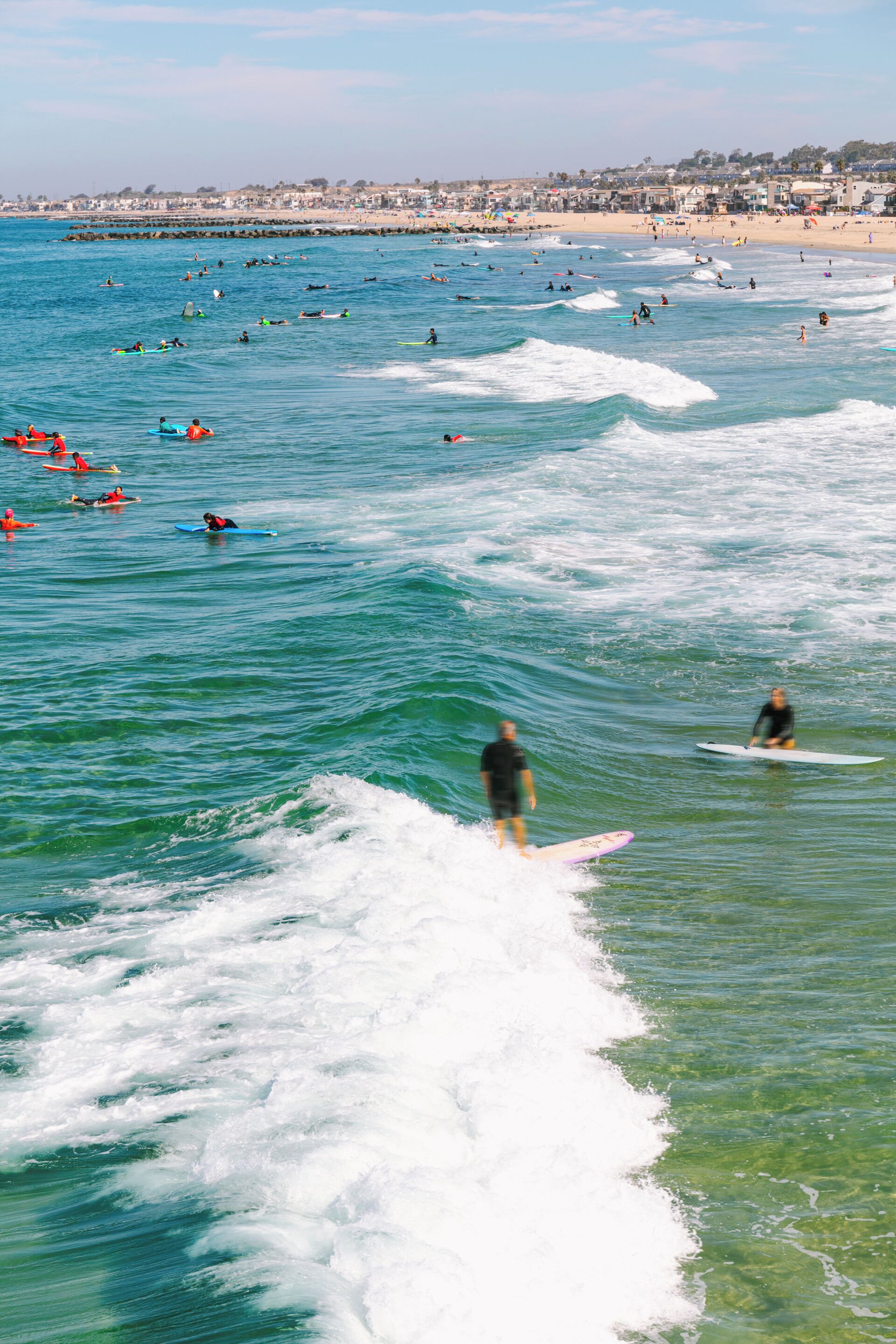 A beach with surfers and swimmers in the water, a pier in the background, and a sandy shore with buildings.