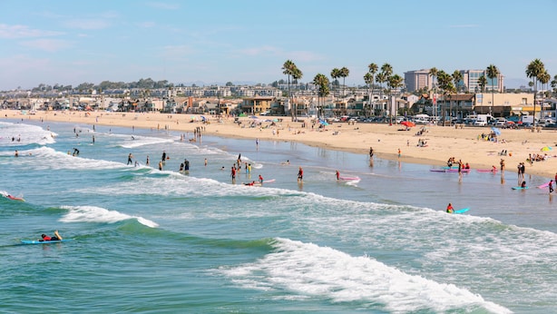 A beach with surfers and swimmers, a sandy shore, and buildings in the background. - Solarena at Newport Beach (Newport Beach)