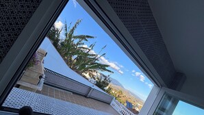 Blick aus einem Fenster auf einen Balkon mit weißer Brüstung, Palmen und eine gebirgige Landschaft.