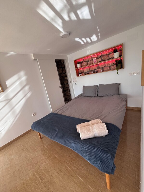 A bedroom with a bed, a wall-mounted shelf with decorative items, a door leading to another room, and a tiled floor.