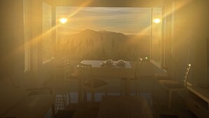Ein Zimmer mit einem großen Fenster und Blick auf eine gebirgige Landschaft. Es gibt einen Tisch mit Stühlen und einen kleinen Tisch mit Gegenständen darauf.
