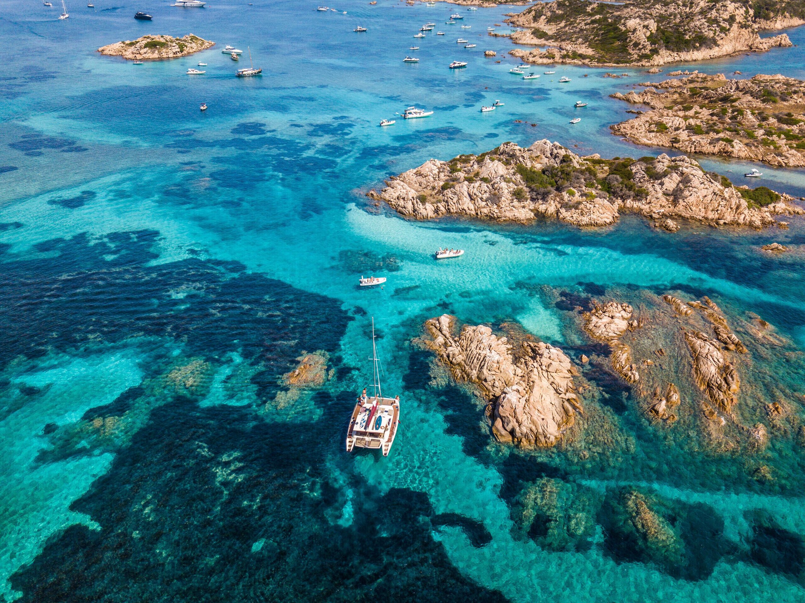 An aerial view of a coastal area with clear turquoise waters, rocky outcrops, and several boats.