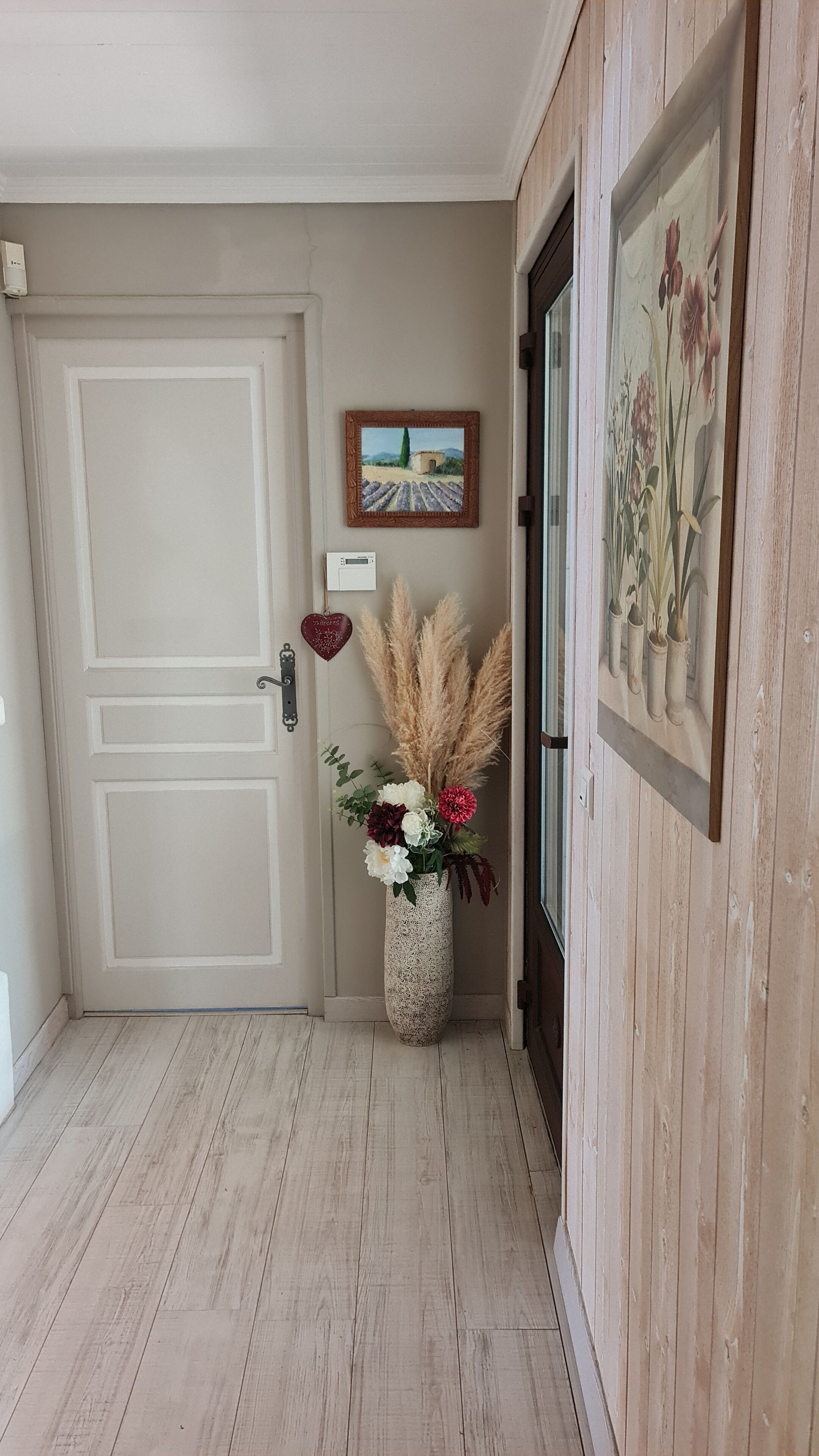 A hallway with a white door, a framed painting, a vase with flowers, and a decorative heart on the wall.