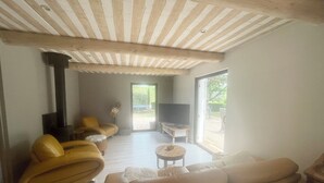 Een ruime woonkamer met een hoge nok, een open haard, een bank, een televisie en een glazen schuifdeur die uitkomt op een buitengebied.