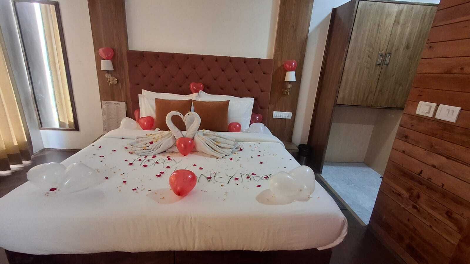 Habitación de hotel con una cama decorada con un arreglo de globos con forma de corazón, un ramo de flores y un letrero que dice 'Feliz aniversario'.