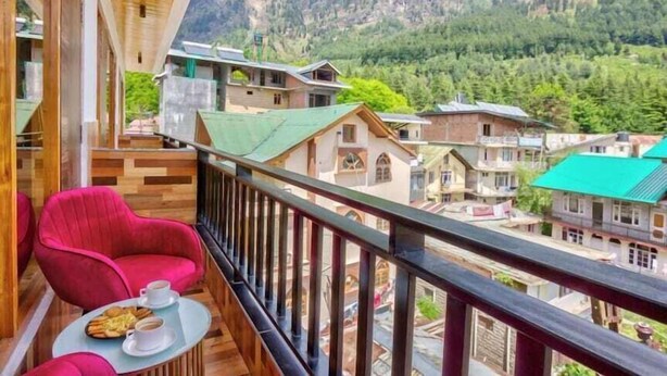 A balcony with a table set for two, overlooking a mountainous village with traditional houses. - Everest Hotel Manali (Manali)