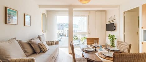 A modern living room with a sofa, dining table, and chairs, a large window with a view of the city, and a hanging light fixture.