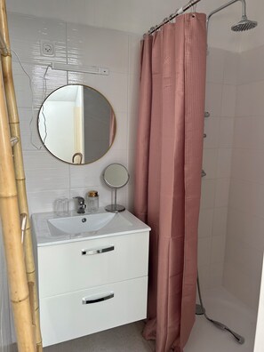 Un baño con tocador blanco, dos espejos, ducha con cortina rosa y una planta de bambú.