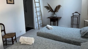 A room with two beds, a chair, a ladder, a vase with dried feathers, and a framed picture on the wall.
