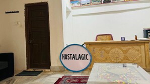 A room with a wooden door, a round sign reading "HOSTALAGIC", a reception desk with a decorative panel, and a wall with framed pictures. - Life Solora Jaisalmer (Jaisalmer)
