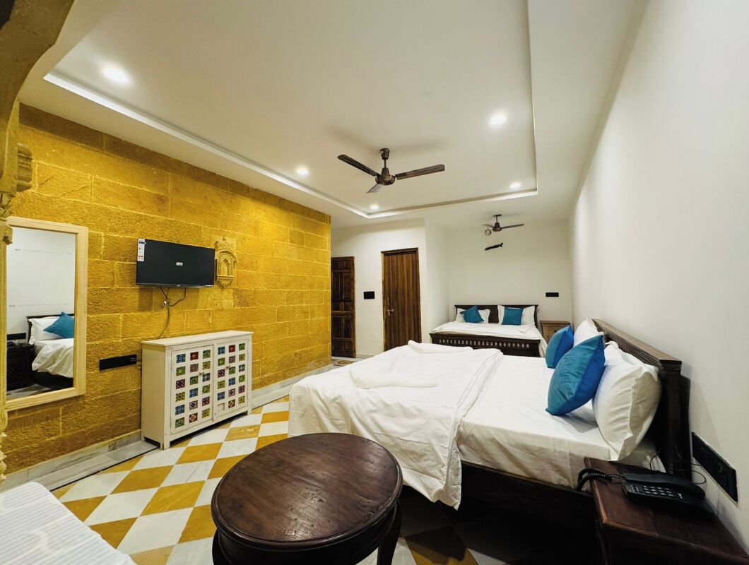 A hotel room with a checkered floor, a bed with white bedding and blue pillows, a wooden nightstand with a telephone, a small round table, a wall-mounted TV, and a ceiling fan.