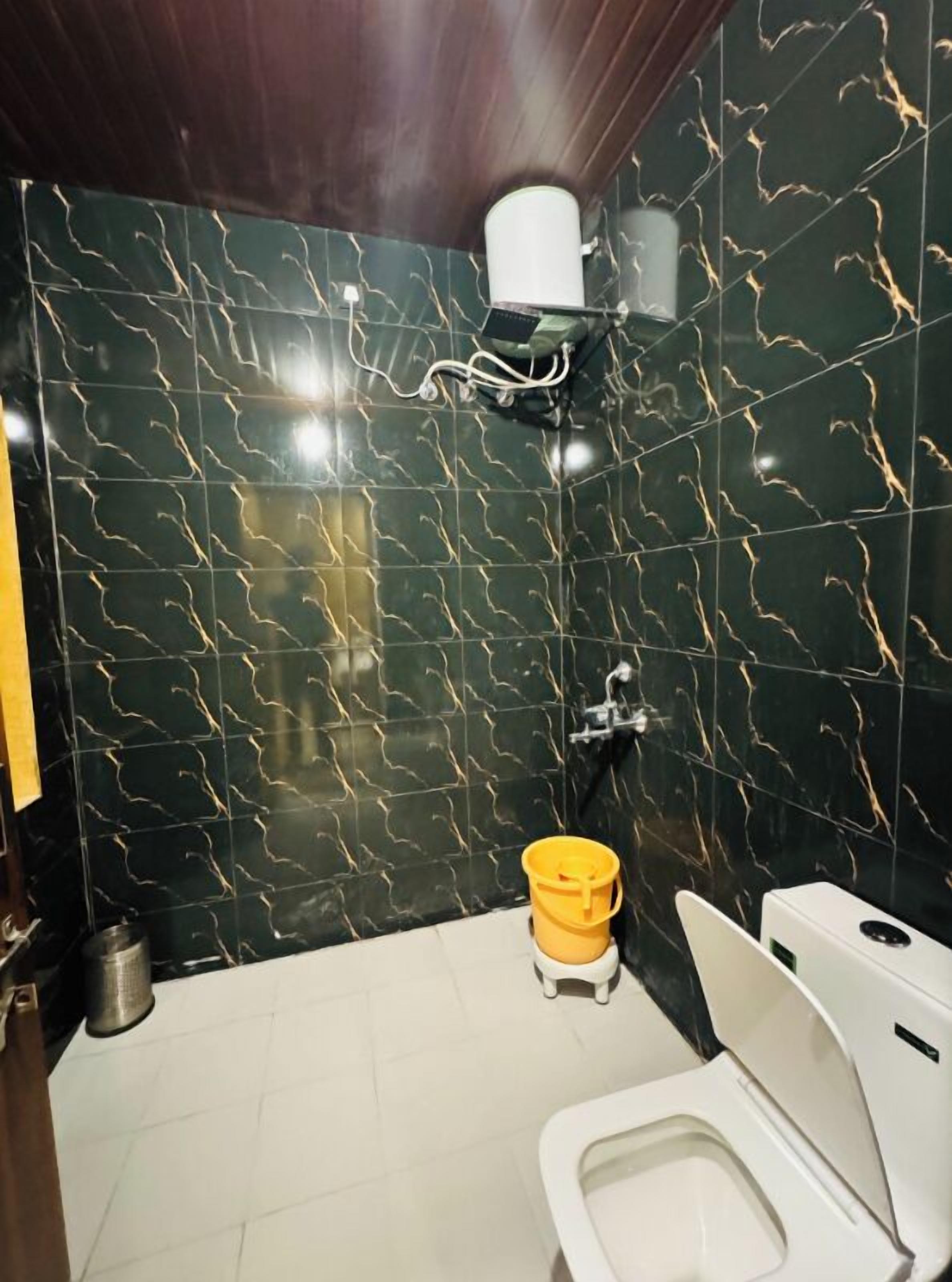 Une salle de bain avec des murs recouverts de marbre foncé, des toilettes blanches, un seau jaune et un sèche-mains blanc.