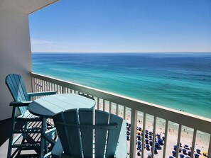 A balcony with two chairs and a table, overlooking a beach with umbrellas and people. - Pelican Beach Top Floor Beachfront with Best Views (Destin)