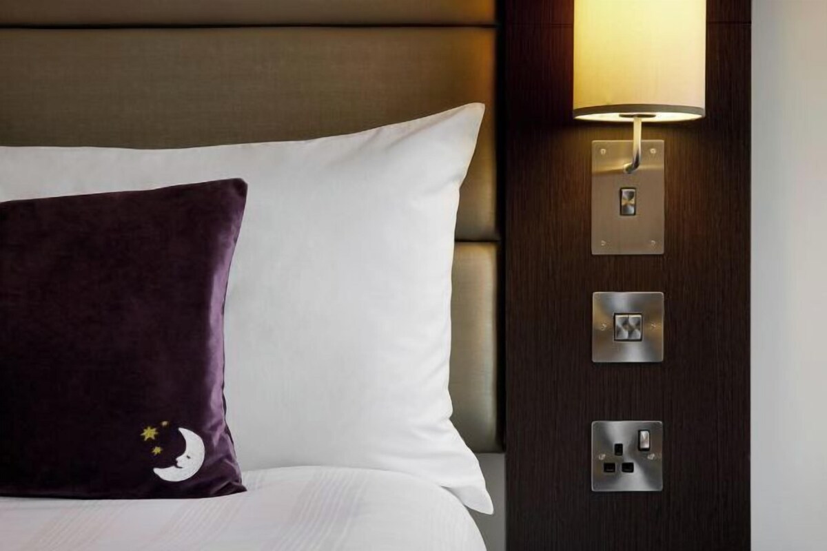A hotel room with a bed, a purple pillow, a white pillow, and a wall-mounted lamp.