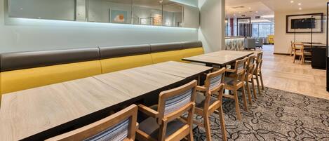 Un espace repas moderne avec de longues tables en bois, des sièges rembourrés et un grand écran fixé au mur.