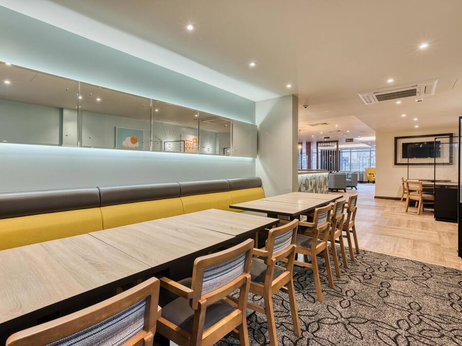 Un comedor moderno con largas mesas de madera, asientos acolchados y una gran pantalla mural.