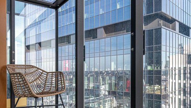 A balcony with a wicker chair, large glass windows, and a city view. - RentPlanet - Apartament Prosta (Warsaw)