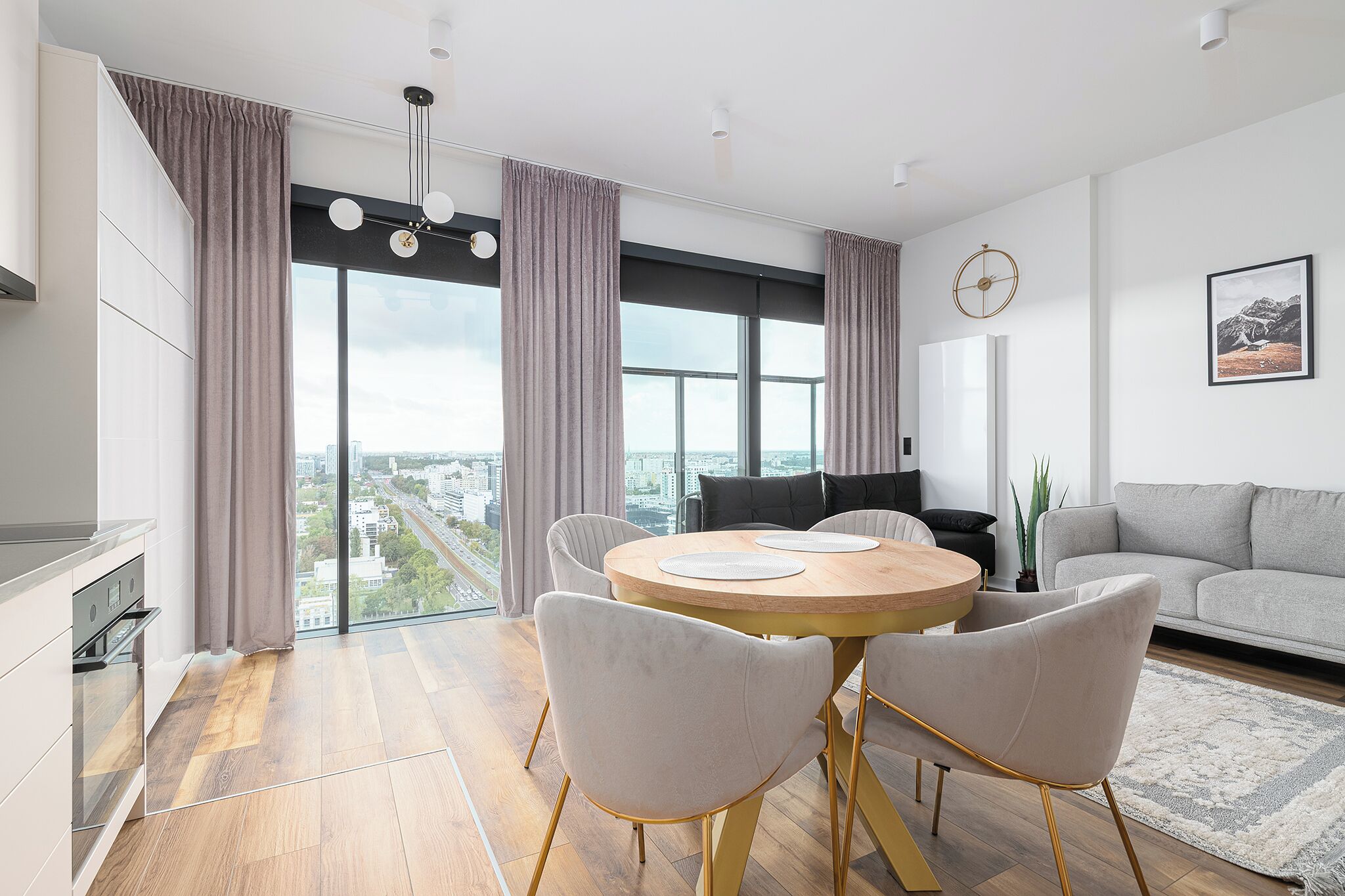 A modern living room with a round dining table, grey sofa, and a large window offering a city view.