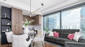 A modern living room with a dining area, a grey sofa with pink cushions, and a large window offering a city view. - RentPlanet - Apartament Prosta (Warsaw)