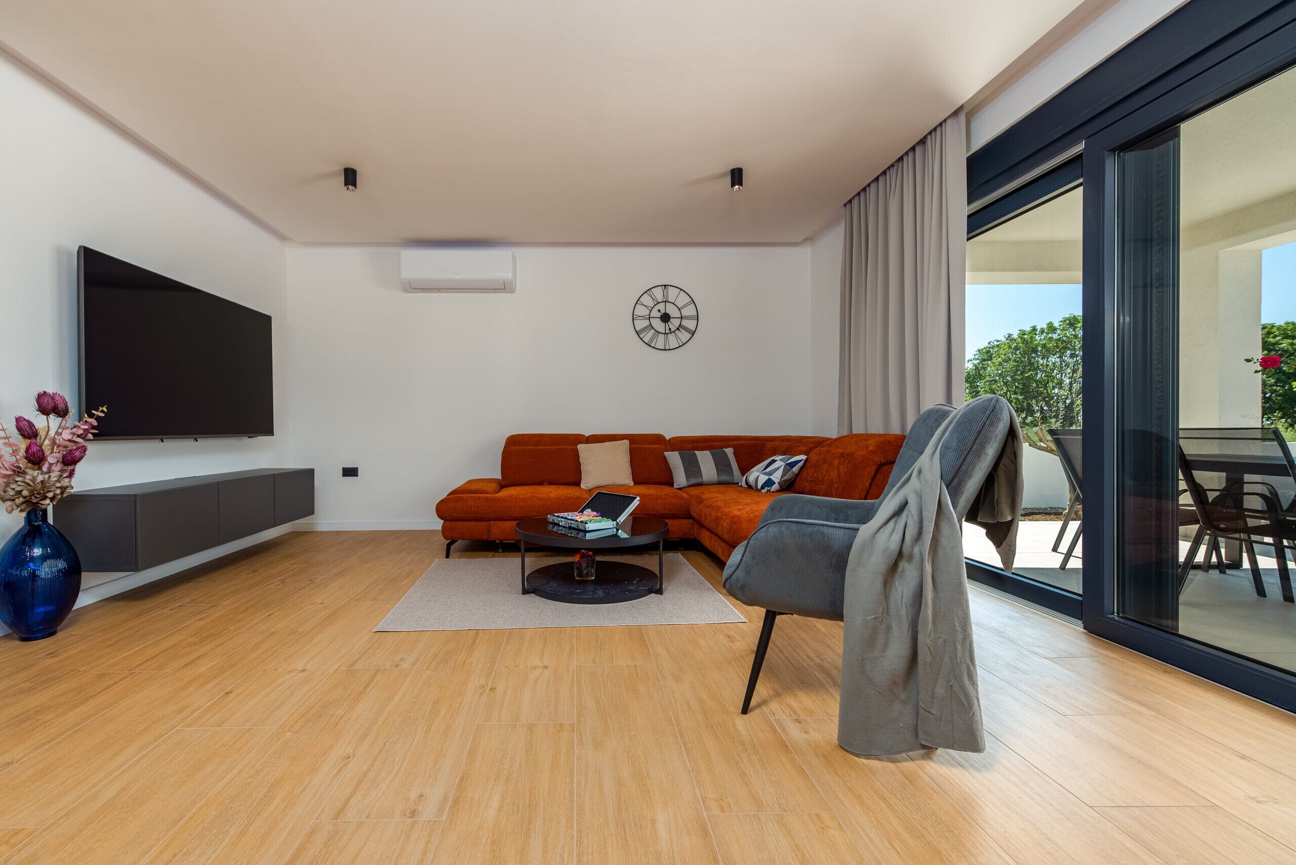 Ein modernes Wohnzimmer mit einem Ecksofa, einem Flachbildfernseher, einem Couchtisch und einem großen Fenster mit Blick ins Freie.