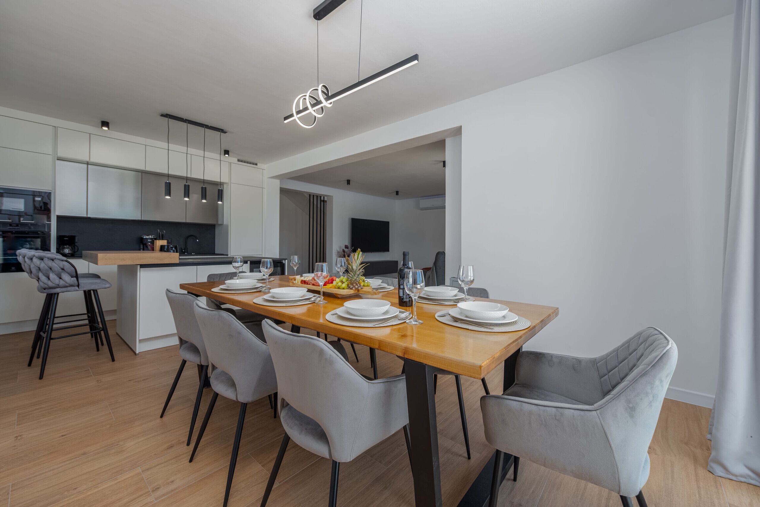 A modern kitchen with white cabinets, a wooden dining table set with plates, glasses, and a fruit bowl.
