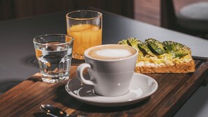 A tray with a glass of water, a glass of orange juice, a cup of coffee, and a slice of avocado toast on a wooden table. - AV366 (Arenas Verdes)