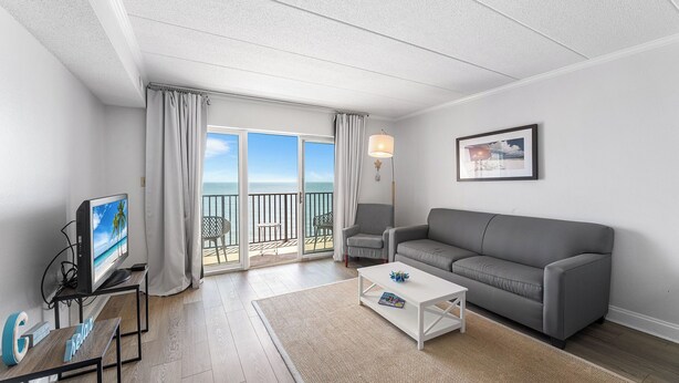 Un salon moderne avec un canapé gris, une table basse, un téléviseur posé sur un meuble et un balcon offrant une vue sur la mer.