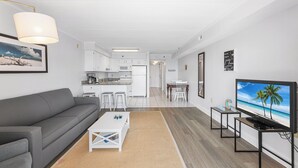 Un salon moderne avec un canapé gris, une table basse, un téléviseur diffusant une scène de plage et une cuisine en arrière-plan.