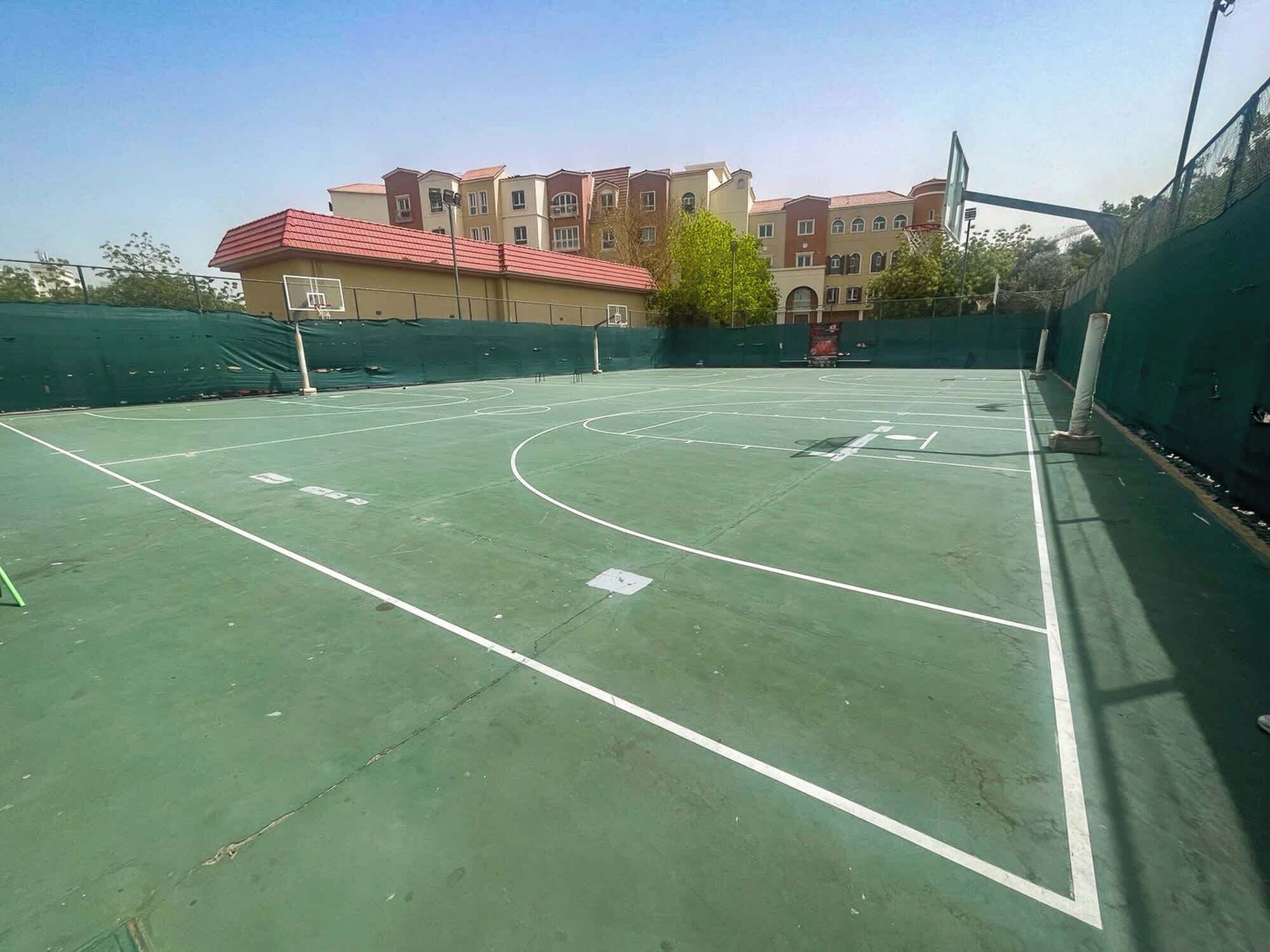 Un terrain de basket en plein air, doté d’une surface verte, entouré d’une clôture verte et avec un bâtiment au toit rouge en arrière-plan.