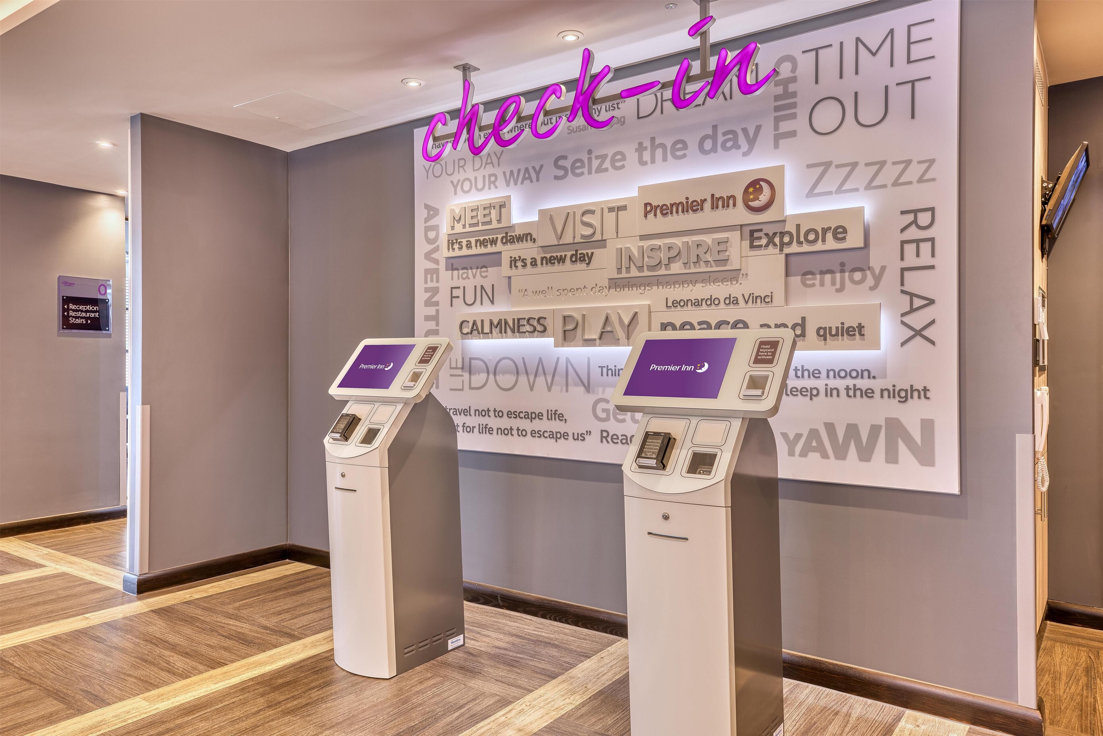 A hotel reception area with a check-in counter, a digital display, and a wall with motivational phrases.