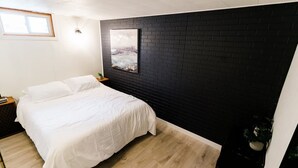 A bedroom with a bed, a nightstand, a wall-mounted light, a window, and a black brick wall.