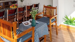 Un coin repas avec des chaises et des tables en bois, un ventilateur au mur et une cuisine en arrière-plan.