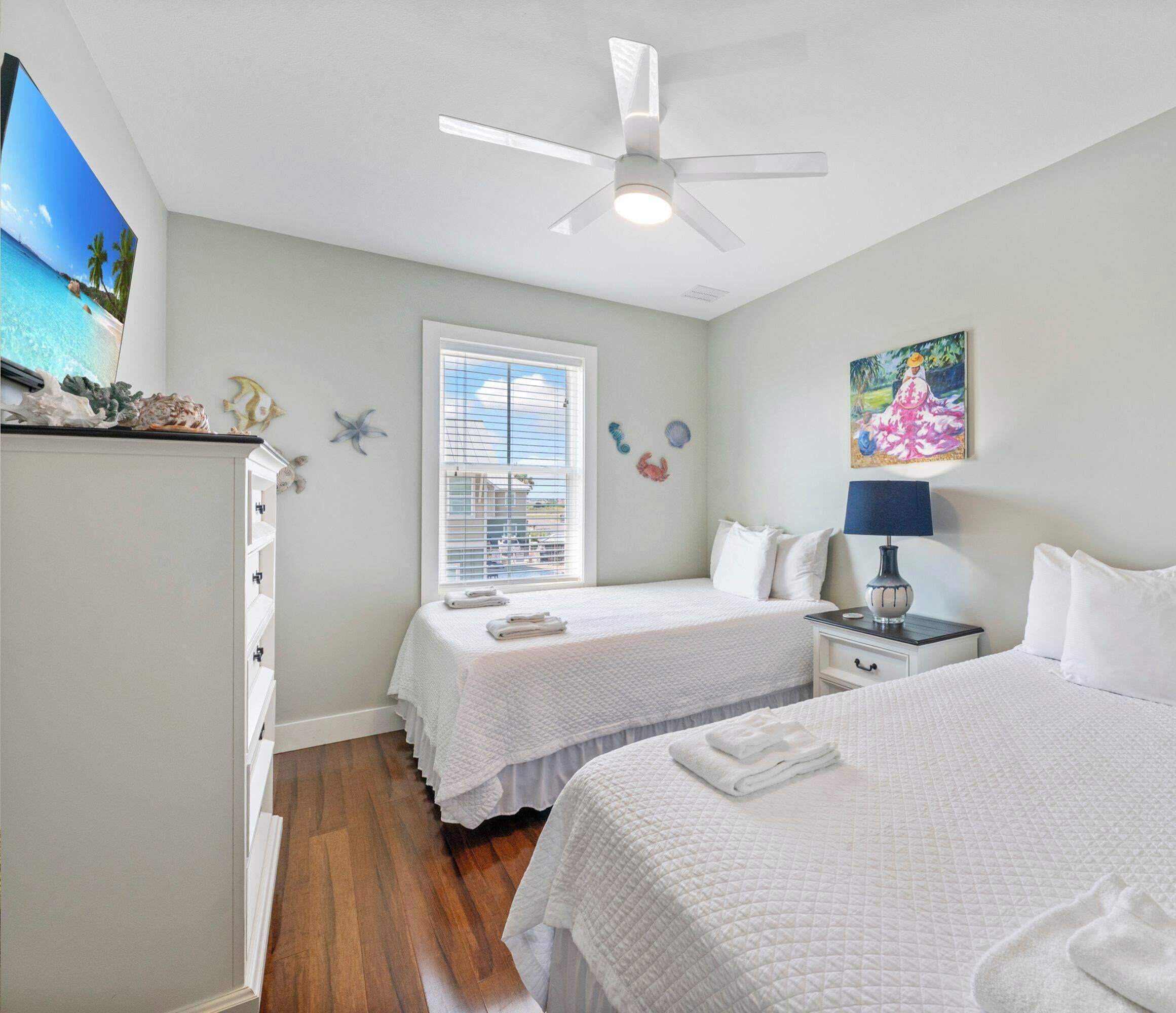 A clean, well-lit bedroom with two beds, a ceiling fan, a window with blinds, and a painting on the wall.