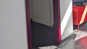 Habitación de hotel con una puerta que da a un pasillo, un reloj en la pared y una mesa roja con una silla negra.