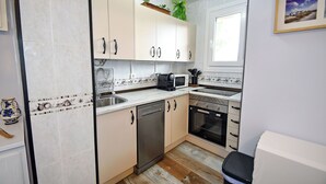 A compact kitchen with a dishwasher, stove, microwave, and sink. There are cabinets with handles and a window above the sink. - Serene Lakeview Retreat (San Martín de Valdeiglesias)