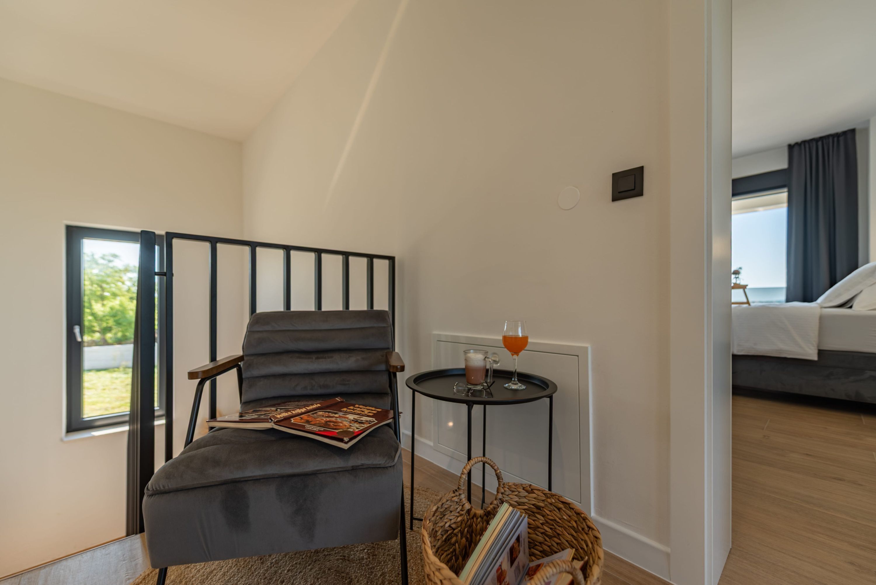 Una habitación moderna con un sillón tapizado gris, una mesita con una bebida y una cesta de mimbre.