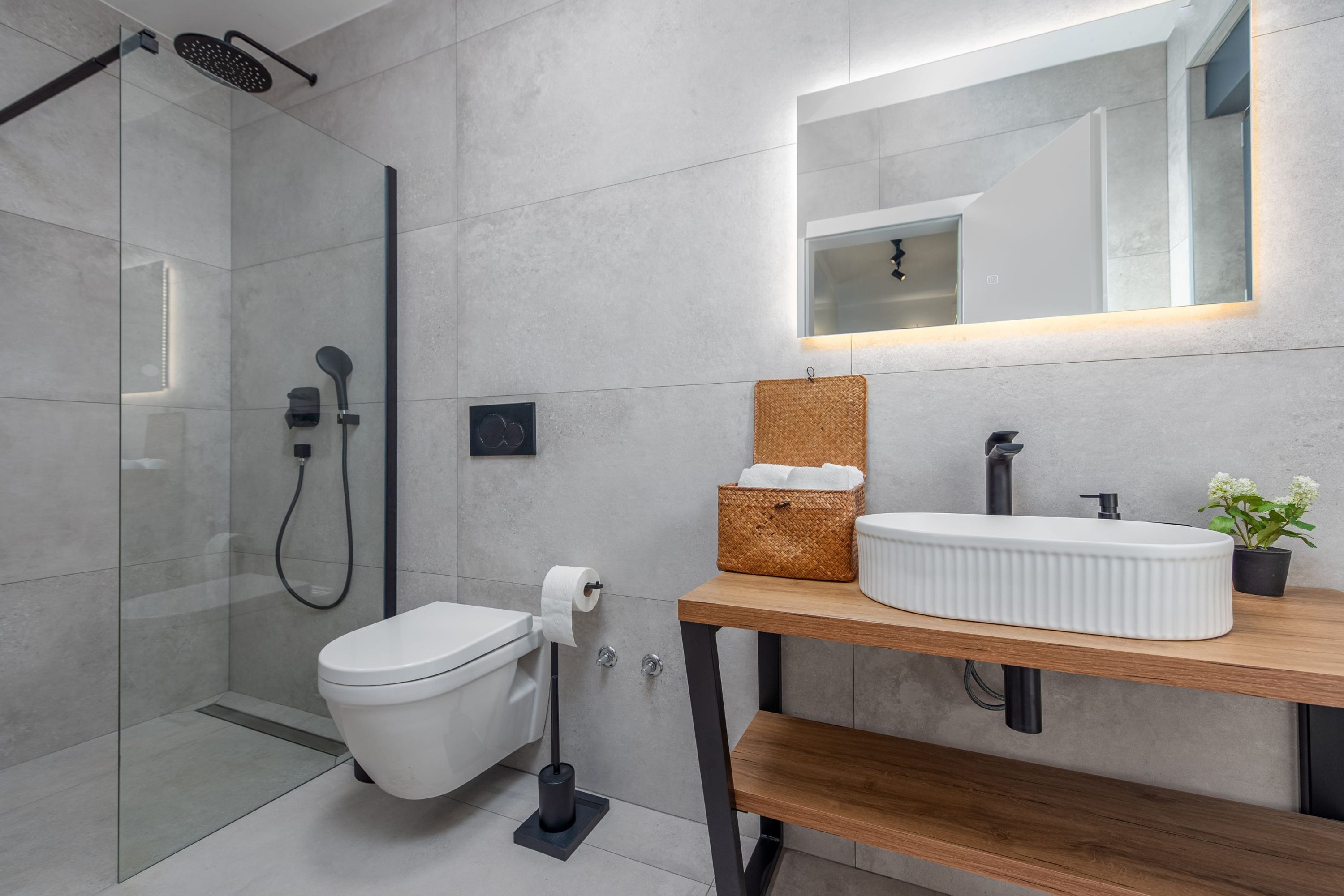 Un baño moderno con ducha envuelta en vidrio, inodoro blanco, tocador de madera con lavamanos blanco y un espejo sobre el tocador.