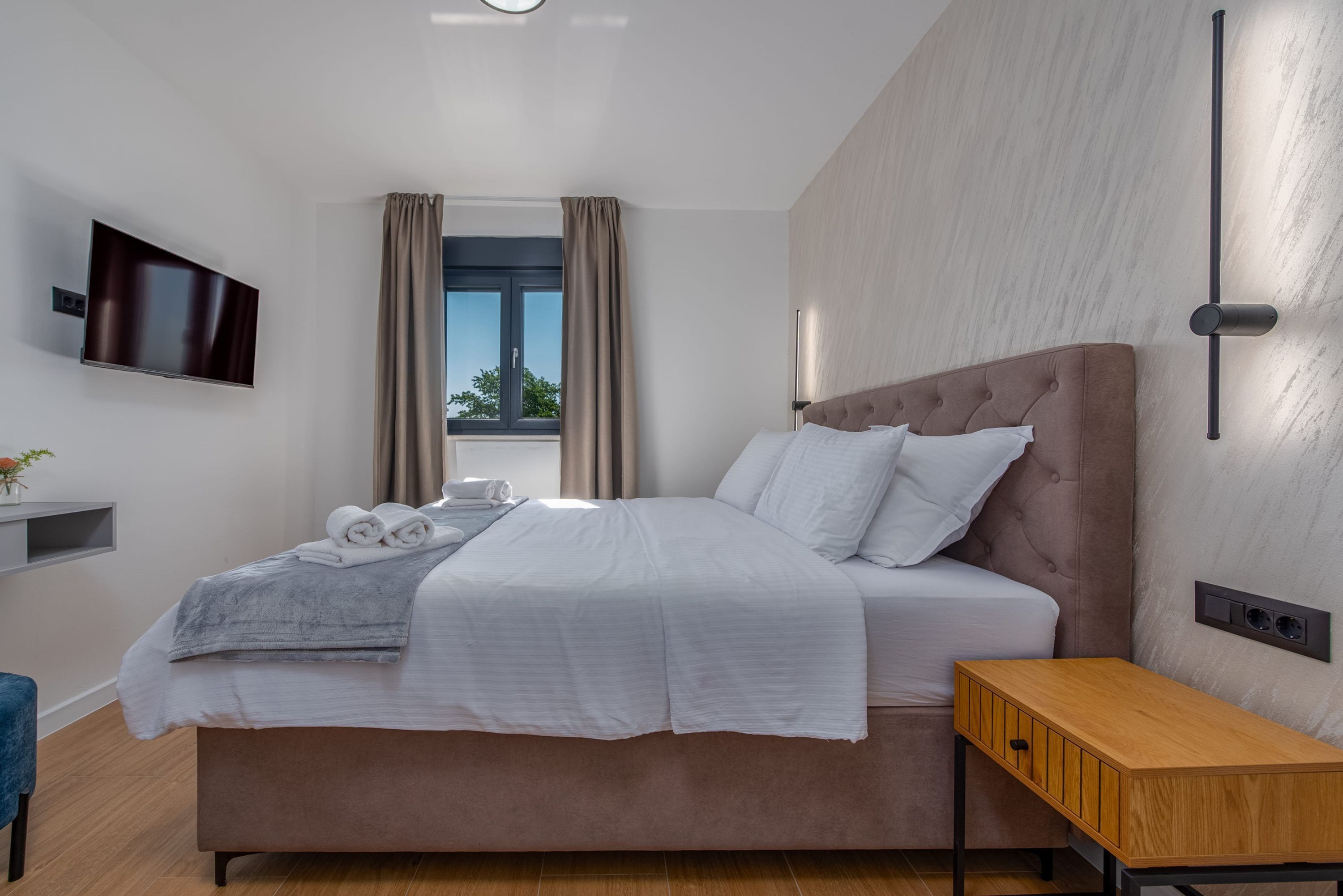 Et moderne hotelværelse med en stor seng, et natbord, et fladskærms-TV på væggen og et vindue med gardiner.